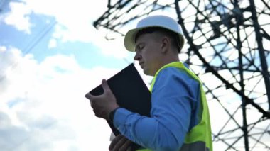 Beyaz kask takan, yansıtıcı yelek giyen ve elinde tablet tutan bir enerji mühendisi elektrik kulesi ve kablolar görüyor. Enerji mühendisi verimli ve güvenli elektrik şebekesi yönetimine odaklanıyor.