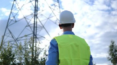 Beyaz bir miğfer, yansıtıcı yelek ve tabletle bir enerji mühendisi elektrik kulesi ve kabloları inceliyor. Enerji mühendisi verimli güç sistemlerini korumaya odaklanıyor.