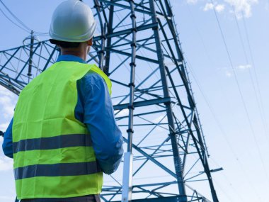 Beyaz kasklı ve yansıtıcı yelekli enerji mühendisi bir elektrik kulesini ve kabloları inceliyor. Enerji mühendisi, elektrik sistemlerinin performansını güvence altına alarak, güvenlik ve verimlilik için altyapıyı değerlendiriyor.