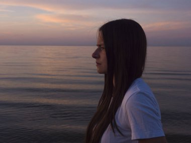 Genç bir kadın, gün batımının deniz kıyısında görülmesinin keyfini çıkarıyor. Gün batımının altın ışıkları sudan yansıyarak büyüleyici ve rahatlatıcı bir atmosfer yaratıyor..