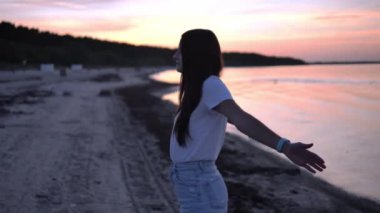 Genç bir kadın deniz kıyısında gün batımına doğru ellerini uzatıyor. Genç kadın uzanırken, güneşin sıcacık parıltısı suya yansıyarak, dikkatli çalışması için sakin bir zemin oluşturur..