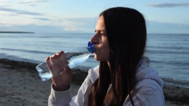 Genç bir esmer kadın, deniz kenarında plastik bir şişeden maden suyu içerek bir anın tadını çıkarıyor. Sahilde otururken maden suyu onu canlandırıyor ve sakin ortamın tadını çıkarıyor..