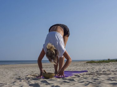 Genç sarışın bir kadın, plajda yoga yaparak kaslarını ısıtıyor. Paspası deniz kenarına yerleştirilmiş. Her esneme hareketinde, açık gökyüzünün altında barışçıl ve odaklanmış bir yoga seansına hazırlanıyor..