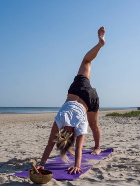 Yoga çalışmasına odaklanmış genç sarışın kadın vücudunu deniz kenarında bir mindere geriyor. Yoga rutini, kumlu sahilde zarifçe akan esneklik ve gücü birleştiriyor. dikey resim