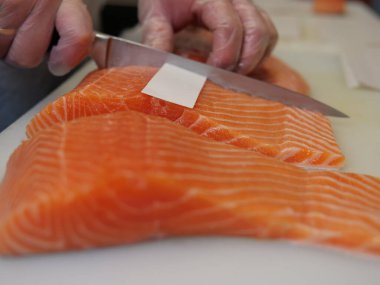 Büyük bir somon balığı, bıçağın canlı renginde ve taze somon dokusuyla ustalıkla dilimlendiği için mutfak tahtasının üzerinde duruyor. Her dilimi kusursuz ve güzel, somonların kalitesini vurguluyor..