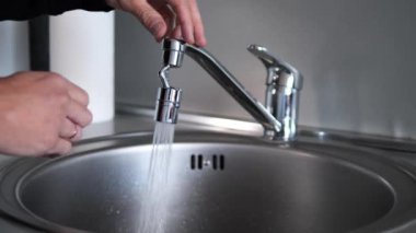 Metal bir musluktan lavaboya doğru sürekli akan su damlalarının her damlası ışığı yansıtıyor. Musluk açık kalıyor, musluktan temiz ve ferahlatıcı bir su akışı geliyor..