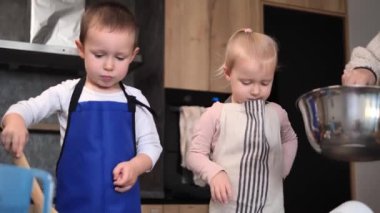 Önlüklü iki çocuk, annelerinin çağdaş bir mutfak ortamında kaseye hamur koymasını hevesle bekleyen tahta kaşıkları tutuyorlar. Hazırlık konsepti Öksürük ve bisküvi
