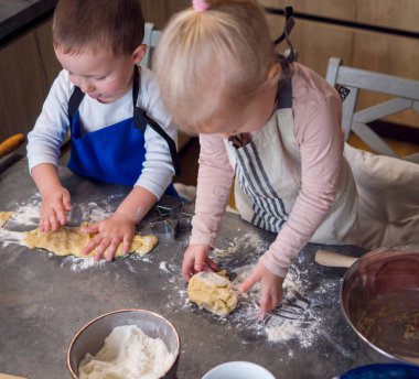 Önlük giymiş iki çocuk, evde kurabiye yapmakla meşguller, mutfak tezgahında hamur yoğurmakla, un, kurabiye keskisi ve kaselerle çevrili.