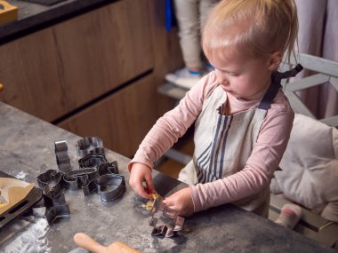 Sarışın yeni yürümeye başlayan çocuk, yeni yıl kurabiyelerini sıcacık mutfakta yapıyor, şekiller yaratmak için hamur kesiciler kullanıyor. Aile geleneğinde pişirmeyi ve eğlenmeyi öğrenmek: neşe, çocukluk