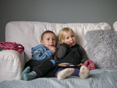 Küçük çocuklar beyaz kanepede oturup televizyon izler ve kucaklarındaki tabaktan atıştırmalıkların tadını çıkarırlar. Sıcak battaniye ve yastıklarla çevrili küçük çocuklar, sıcak ve rahat bir ortam yaratıyor.. 