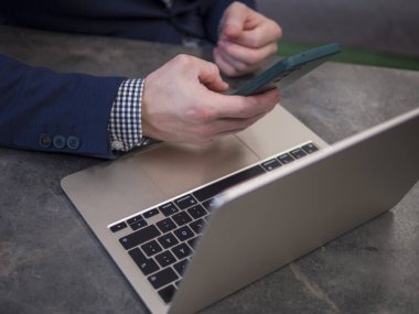 Mavi takım elbiseli ve kareli gömlekli bir iş adamı, akıllı telefon ve dizüstü bilgisayar kullanıyor. İşadamı bilgisayar teknolojisi üzerine uzaktan çalışma görevleri ve iletişim kavramına odaklanıyor 