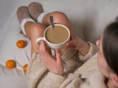 Sıcak bornoz ve sıcak çoraplar giymiş genç bir kadın yatağa oturup sütlü kahvenin tadını çıkarıyor. Kahve fincanı ile rahatlarken mandalinalar ona dokunuyor. Sabah barışı ve kahvesinin tadını çıkarıyor.. 
