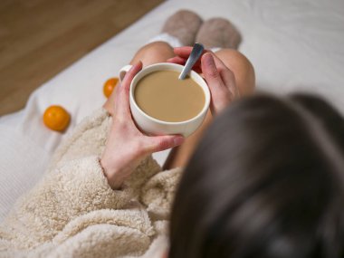 Bornozlu ve rahat çoraplı genç bir kadın sıcak bir fincan kahveyle huzurlu bir sabahın tadını çıkarıyor. Mandarinler, rahatlatıcı kahve fincanıyla rahatlarken yakınındaki sahneyi büyütür. 