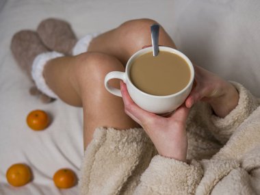 Bej bornozlu ve tüylü terlikli bir kadın elinde sütlü sıcak bir fincan kahveyle yatakta oturuyor. Yakınındaki mandalinalar sabah kahvesini yudumlarken rahat bir dokunuş ekliyor. 