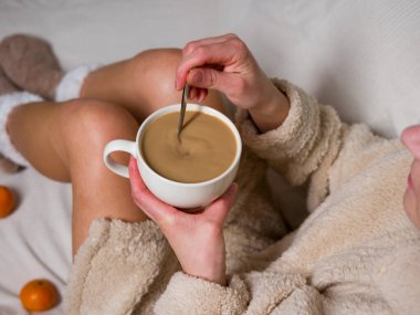 Sıcak bornozlu kadın yatakta rahatlıyor, bir fincan sıcak kahvenin tadını çıkarıyor ve yavaşça karıştırıyor. Yakınlardaki taze mandalinalar huzurlu bir atmosferi canlandırıyor ve bir fincan kahveyle huzurlu bir sabah geçiriyor.