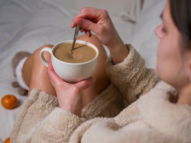 Rahat bornozlu bir kadın yumuşak beyaz bir battaniyeye oturur ve nazikçe kahvesini karıştırır. Sıcak çorap giyiyordu, bir fincan kahvenin tadını çıkarıyordu, bir fincan kahveyle sakinlik yaratıyordu. Yakınlarda mandalina var.