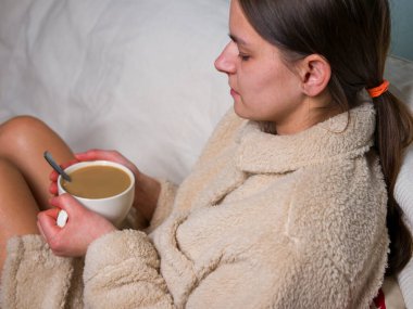 Rahat bir bornoz giymiş genç bir kadın, kanepede rahatça oturuyor, elinde bir fincan kahveyle sıcak bir fincan kahvenin tadını çıkarıyor, evinde rahatlama konseptini kucaklıyor.