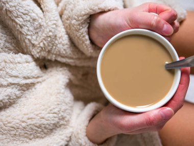 Kabarık beyaz bornozlu kadın sıcak kahve ve sütle dolu beyaz bir fincan tutuyor. Kahvesini rahat bir evde içmeyi seviyor, kahveyle rahatlığı kucaklıyor. Ev rahatlığı ve sıcaklığı kavramı. 