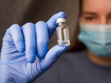 Cerrahi maske takan doktor ve mavi lateks eldivenler temiz sıvı içeren tıbbi bir şişe tutuyor, tıbbi prosedüre dikkatlice hazırlanıyor, tıbbi şişeyle güvenliği vurguluyor.. 