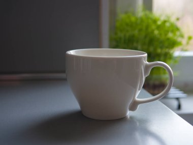 Gri bir mutfak tezgahında dinlenen büyük beyaz kupa, bir pencerenin yanında yeşil bir bitki, rahat ve ferahlatıcı bir atmosfer yetiştiriyor. Sabah anları için mükemmel.