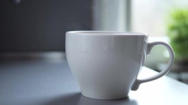 Yumuşak odak penceresinin yanındaki beyaz seramik kahve fincanından yavaşça yükselen sıcak buhar bulanık yeşil bitki ile sabah mutfağında huzurlu bir atmosfer yaratıyor.