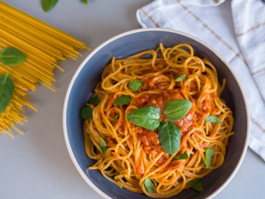 Taze fesleğen yaprakları ve zengin domates sosuyla mavi bir kasede servis edilen Balanesi makarnası yanında beyaz çizgili bir peçeteyle birlikte çiğ spagetti ve fesleğen yaprakları.