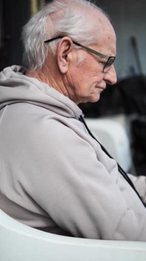 Profilde oturan yaşlı bir adam, yumuşak ışıkta kadehleri nazikçe kaldırıyor. Gri kapüşonlu yaşlı adam sakin atmosfere yansıyor. Yaşlı adam huzurlu bir düşünce anının tadını çıkarıyor. Dikey video