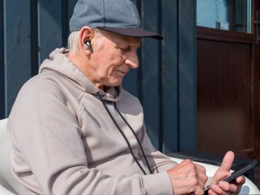 Sunny Garden 'da kablosuz kulaklıklı akıllı telefon kullanarak dışarıda rahatlıyor. Kıdemli adam sakin dijital anın tadını çıkarıyor. Kıdemli adam barışçıl teknoloji odaklı yaşam tarzını benimsiyor.. 