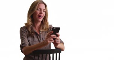 Beyaz arka planda oturan, akıllı telefondan resimlere bakan orta yaşlı beyaz bir kadın. Stüdyoda cep telefonuyla fotokopi çekilen kadın fotoğrafları. 4k