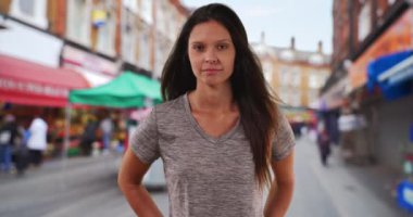 Millennial kadınının portresi ticari bölgedeki caddede kameraya bakıyor. Şehir merkezindeki kaldırımda rüzgarda uçuşan saçlarıyla dikilen günlük elbiseli bir kız resmi. 4k