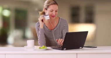 Beyaz kadın mutfak tezgahında oturuyor ve kredi kartıyla laptop 'dan alışveriş yapıyor. Evde bilgisayarla gülümseyen kadın alışverişi. 4k