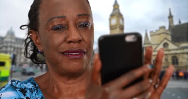 笑顔の黒人女性はスマートフォンで孫の写真を見ている 高齢の女性はロンドンの通りで携帯電話を使う — ストック動画