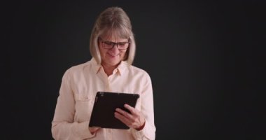 Saf gri arka planda taşınabilir tablet bilgisayarla oynayan mutlu yaşlı kadın. Neşeli kahkahalar atan son sınıf kadını gri arka planda ped kullanmaktan zevk alıyor. 4k