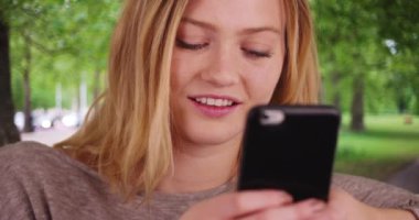 20 'li yaşlarda beyaz bir kız parkta akıllı telefonuyla mesajlaşıyor. Gülümseyerek gezen mutlu ve çekici bir kadın. 4k