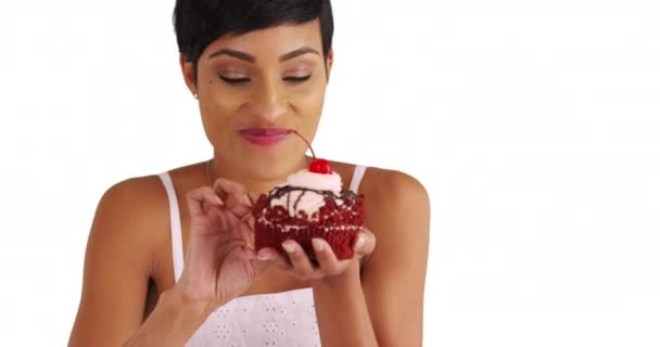 コピースペース付きの白い背景に大きな赤いベルベットのカップケーキを食べる魅力的な黒人女性のクローズアップ 20代の女性のポートレートは スタジオでコピースペースで笑顔で空想的なデザートを楽しんでいます — ストック動画