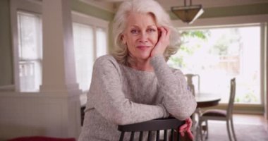 Evdeki kameraya gülümseyen yaşlı bir kadının portresi. 50 'li yaşlarındaki çekici olgun bir kadın kameraya gülümsüyor elleri kapalı bir yerde dinleniyor. 4k