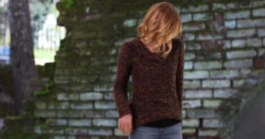 Açık havada tuğla zemine karşı duran beyaz bir kadın. Gülümseyen, kısa sarı saçlı, süveter giyen, güz gününde elleri ceplerinde duran bir kadın. 4k