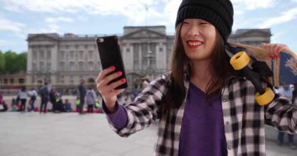 幸せなヒップ100万人の女性がロンドンの技術電話カメラで自撮りします ヨーロッパでスケートボードを保持する若いアジアの女性の近景 — ストック動画