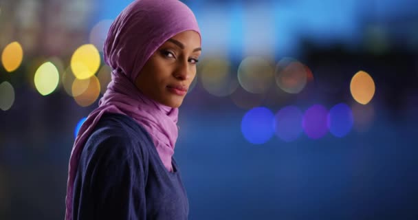 穿着紫色头巾的黑人女性形象严肃地看着相机 穆斯林妇女晚上站在城市里 手握漂亮的蓝色防波灯 — 图库视频影像
