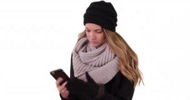 Hoş şapkalı ve fularlı genç çekici bir kadın beyaz arka planda mobil cihazla mesajlaşıyor. Stüdyoda kopyalanmış akıllı telefon kullanmak için kadın eldivenlerini çıkarıyor. 4k