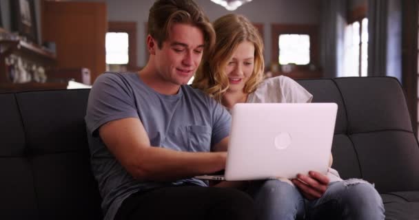 魅力的な白人男性と女性は ラップトップ上のリビングルームのブラウジングビデオでソファに座って 家庭内のコンピュータを使用して幸せな若いカップル 笑顔と笑い — ストック動画