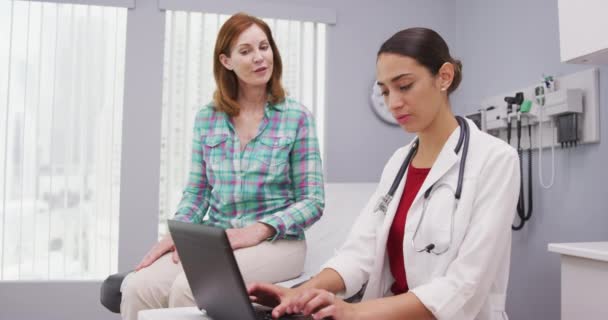 診療所内で高齢者と話をしながらノートパソコンを使った若いラテン系医師のポート 中年患者の終わり若い女性医師の健康上の問題と議論 — ストック動画
