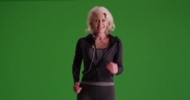 Yaşlı kadın yeşil ekranda kulaklıklarıyla kameraya doğru mutlu bir şekilde yürüyor. Yeşil ekranda spor kıyafetleri içinde koşan yaşlı bir kadın anahtarlanmak ya da birleştirilmek için.