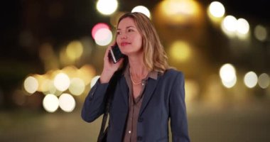 Orta yaşlı, beyaz bir kadın gece şehir merkezinde cep telefonu görüşmesi yapıyor. Güler yüzlü, gülümseyen profesyonel bir kadın akşam sokakta telefon görüşmesi yapıyor. 4k