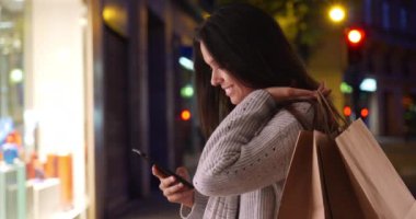 Alışveriş torbalı Millennial kadını gece sokakta telefon özçekimi çekiyor. Gri kazaklı güzel bir kadın akıllı telefonuyla fotoğraf çekiyor. 4k