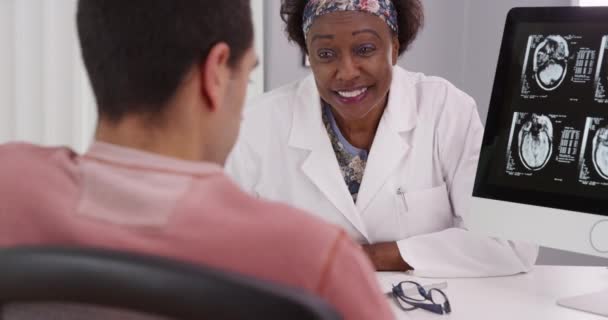 中年黑人医生向年轻男性病人咨询有关颅骨扫描的问题 年轻的男性病人在医生办公室的电脑上扫描他的大脑 — 图库视频影像