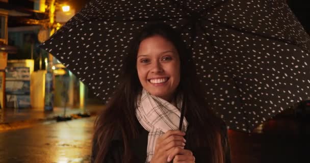 雨の中 夜の街中の美しい女性が水玉の傘を持っている 黒いレインコートの回転傘と笑顔で白人の千年紀の女性 — ストック動画