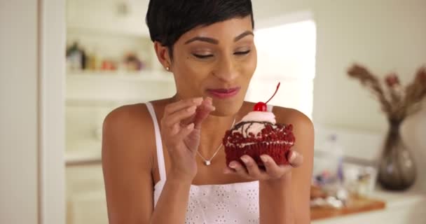 魅力的な黒の女性がお食事を楽しんで閉じます キッチン内のカップケーキの霜を試飲 20代の女性の肖像画は 大きな赤いベルベットのカップケーキを保存し マラッシーノの桜を吸います — ストック動画