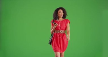 Yeşil ekran arka planında yürüyen ve akıllı telefon müziği dinleyen mutlu siyahi kadın. Şık genç bayan yeşil ekranda şarkı söylemek için telefonu kullanırken kameraya doğru yürüyor. 4k