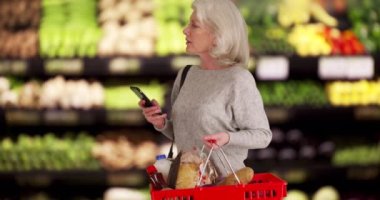 Üretim reyonunda alışveriş yapan Kafkasyalı üst düzey kadın telefonla mesajlaşırken eşyalara bakıyor. Marketten alışveriş yapan yaşlı bir kadın sepeti tutuyor. 4k